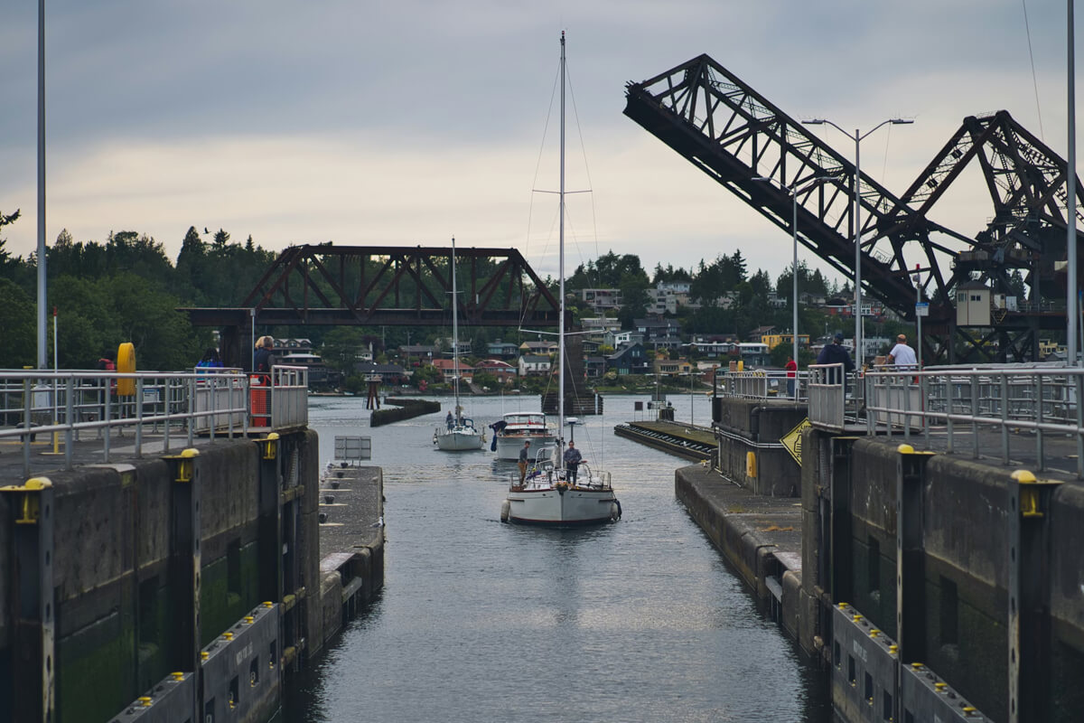 Boats passing through the Ballard Locks in Seattle with the drawbridge raised and people watching from the walkways on a cloudy day.