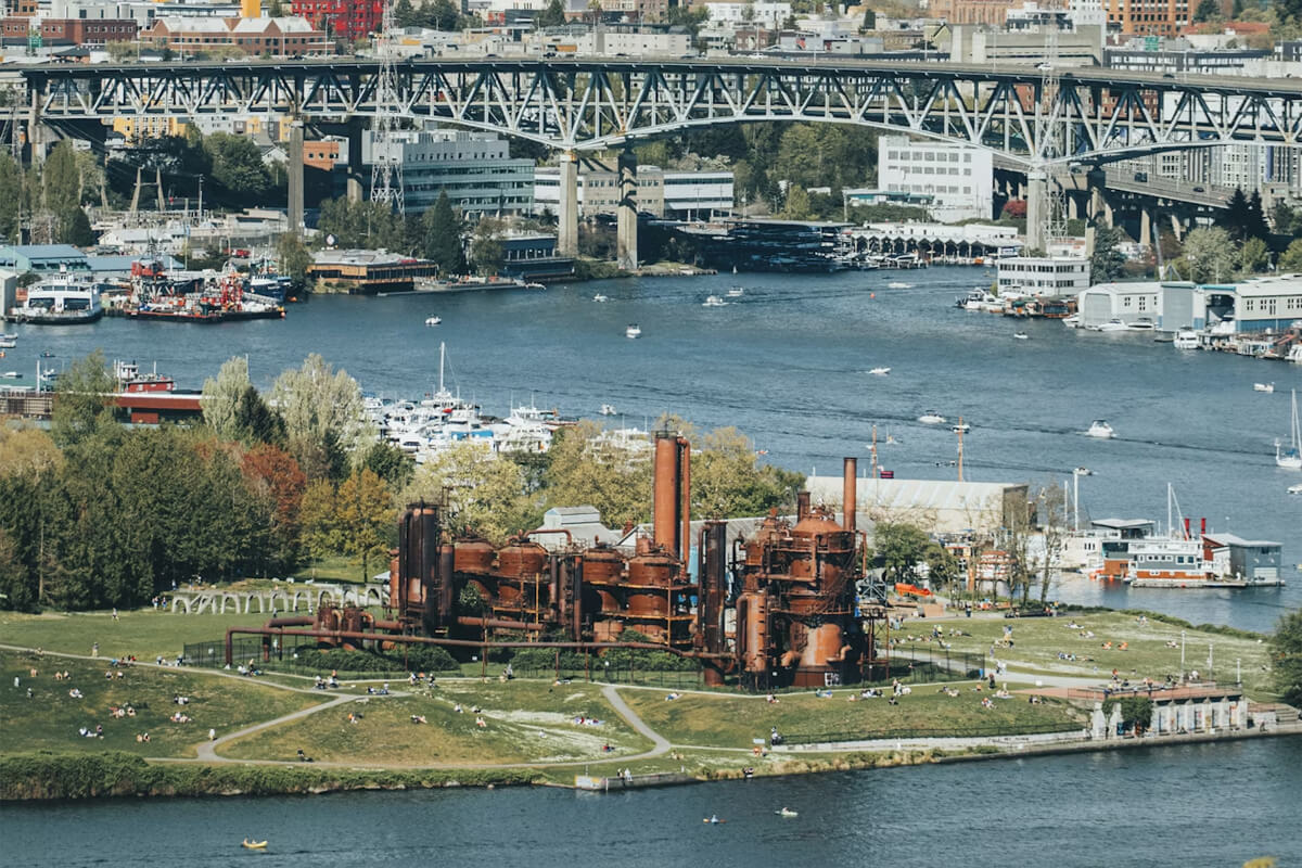 View of Gas Works Park on Lake Union in Seattle, with the historic gas plant structures, people relaxing on the grass, and boats on the water beneath the Fremont Bridge.