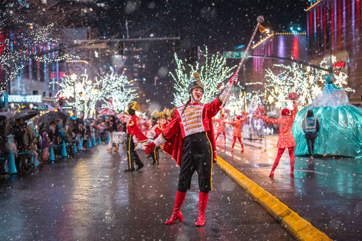 Performer in a toy-soldier costume leading the Snowflake Lane holiday parade in Bellevue, surrounded by lights, dancers, and falling snow.