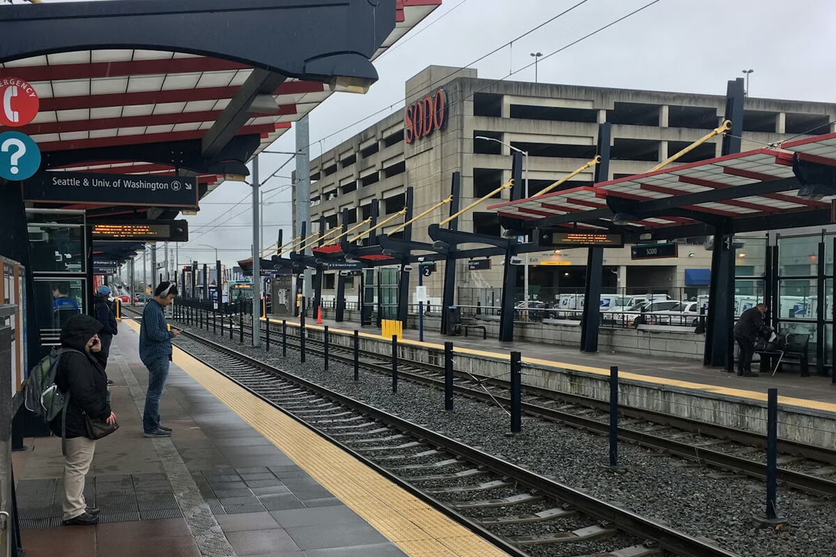 Passengers waiting on the Sound Transit Link light rail platform, with signage toward Seattle and the University of Washington.