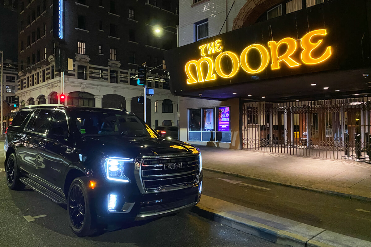 Starline’s black GMC Yukon parked curbside at night in downtown Seattle, with headlights on and “The Moore” marquee glowing above the entrance.