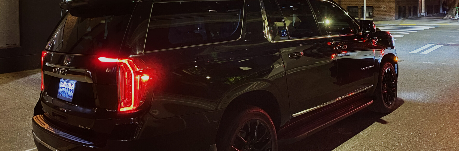 Starline's black GMC Yukon SUV parked curbside on a city street at night, with red taillights glowing and streetlights reflecting on the glossy paint.