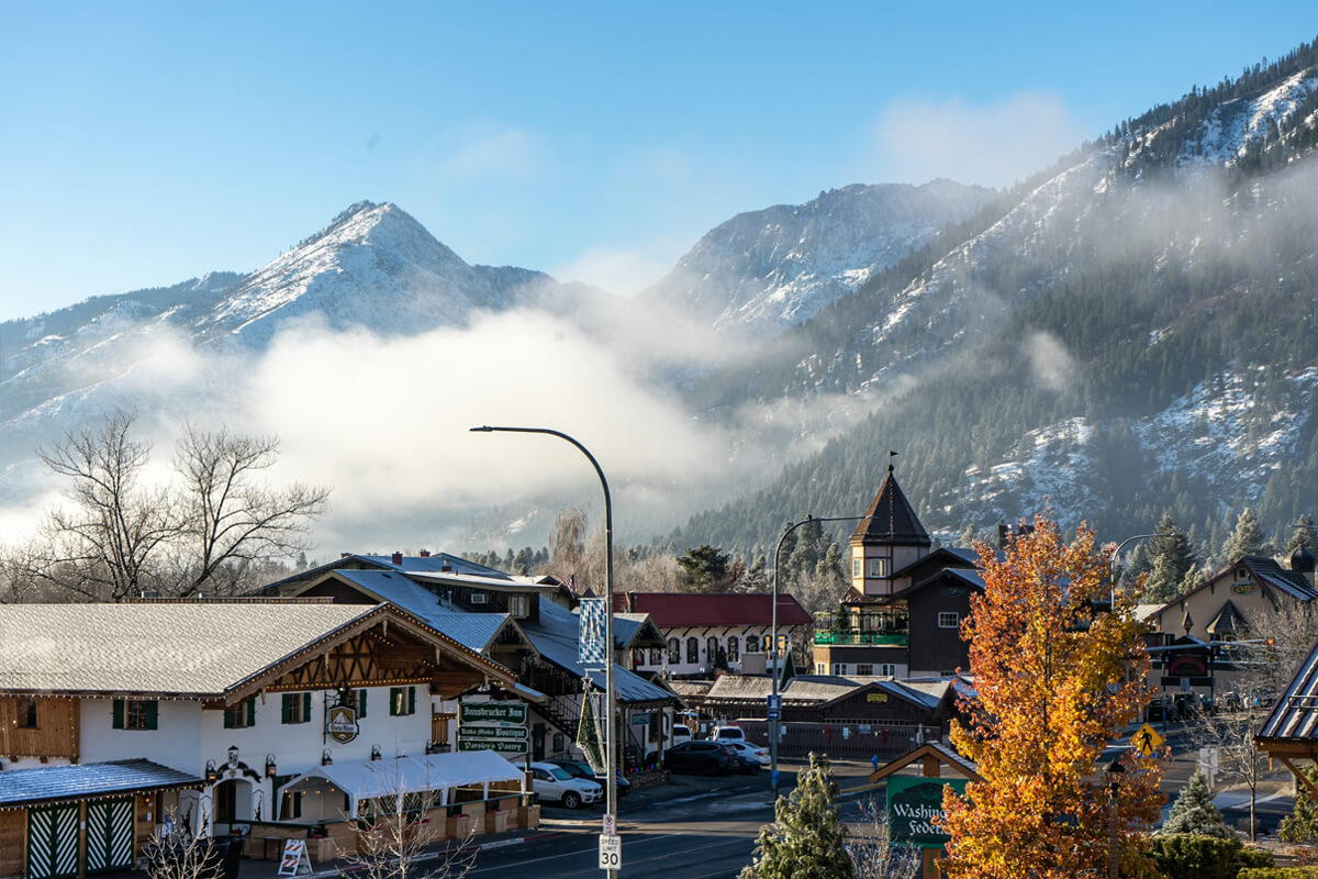 Wide scenic view of downtown Leavenworth with alpine-style buildings, fall color, and snow-dusted mountains rising behind the village
