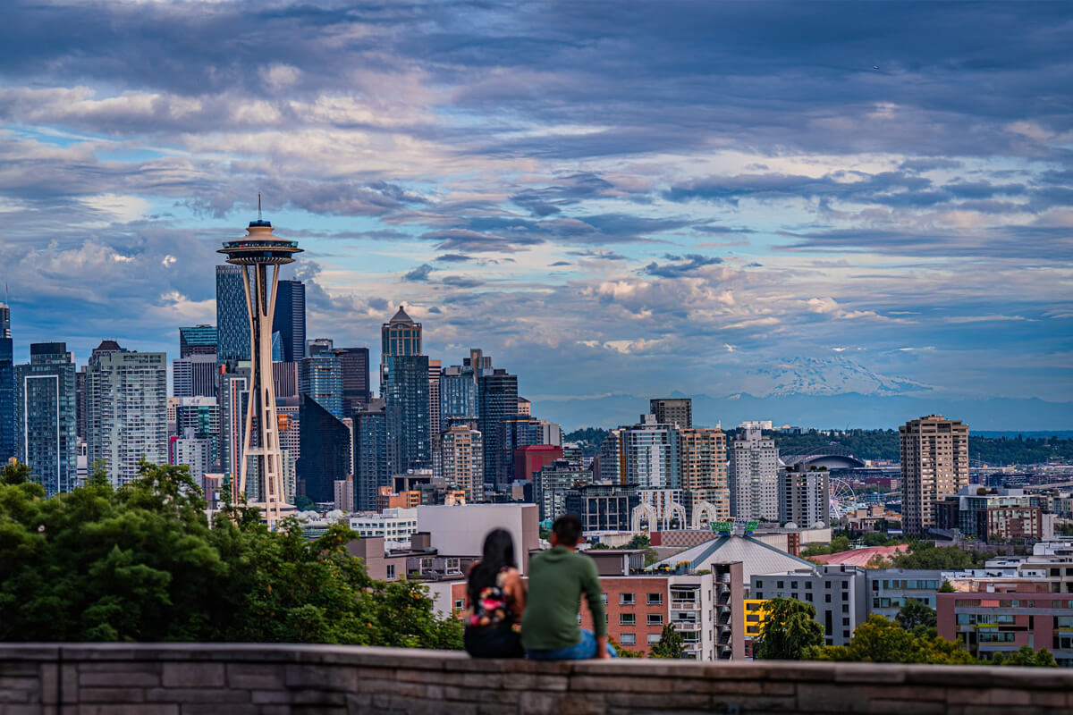 Couple enjoying a panoramic view of the Seattle skyline and Space Needle from Kerry Park at sunset, with Mount Rainier visible in the distance.