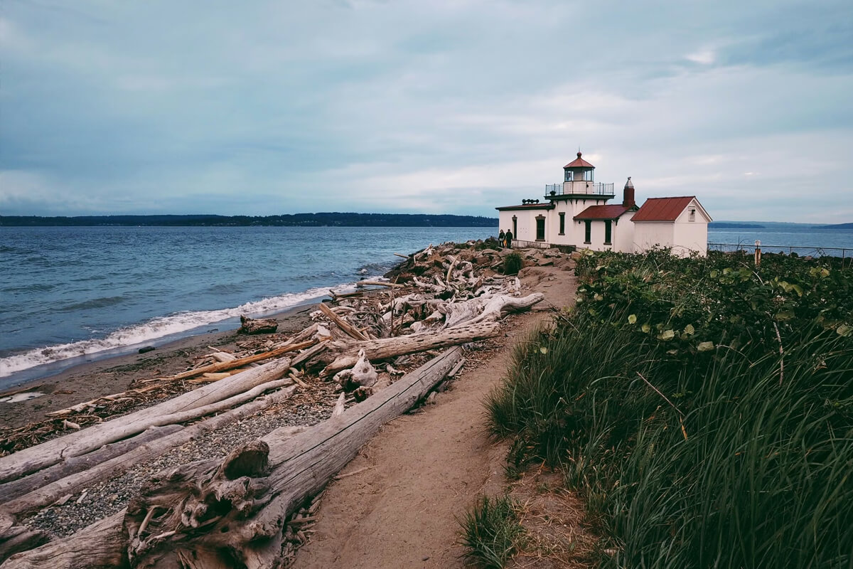 West Point Lighthouse at Discovery Park in Seattle, with driftwood along the beach and waves coming in on a cloudy day.